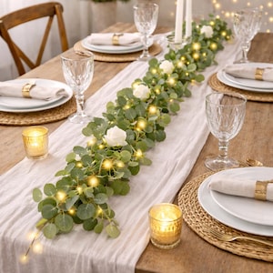 May include: A beautifully set dining table with a white linen runner, adorned with a garland of eucalyptus leaves, white roses, and warm fairy lights. The table setting includes plates, napkins, gold napkin rings, and wine glasses.