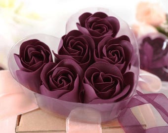 May include: A heart-shaped clear plastic container filled with six deep purple rose-shaped soaps. The container sits atop a cardboard box and is tied with a light pink ribbon. A second container with roses is visible in the background.