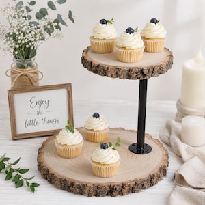 May include: A two-tiered cupcake stand featuring two circular wood slices with natural bark edges connected by a black metal pole. Four cupcakes with white frosting and single blueberries are arranged on the tiers. A sign reads Enjoy the little things.