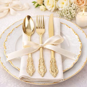 May include: A formal place setting featuring gold-colored flatware: a spoon, fork, and knife, resting on a white napkin tied with a cream-colored ribbon. The plates are white with a gold trim. A candle and flowers are in the background.