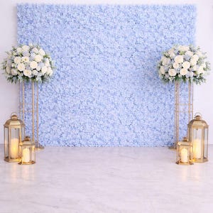 May include: A light blue floral wall backdrop with two floral arrangements on gold stands. Gold lanterns with lit candles are in front of the stands. The floral arrangements are white and light blue.