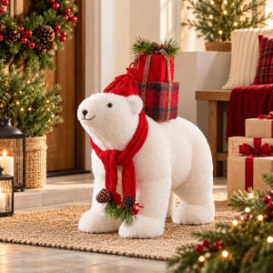 May include: A plush white polar bear figurine wearing a red knit hat and a matching red scarf adorned with small pinecones and greenery. The bear carries two stacked gift boxes on its back, one solid red and one red and green plaid.