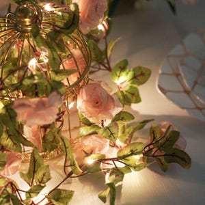 May include: A decorative gold birdcage is adorned with pink roses and green leaves, illuminated by small fairy lights. The floral arrangement is part of a garland, creating a romantic and elegant ambiance. The birdcage is approximately 8 inches tall.