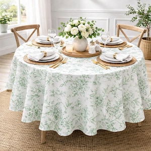 May include: A round dining table set for a meal. The table is covered with a white tablecloth featuring a green floral pattern. Each place setting includes white plates, a napkin, and gold-colored cutlery. A vase of white flowers sits in the center.