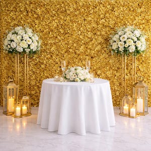 May include: A round table with a white tablecloth features a central white floral arrangement, two champagne flutes, and small votive candles. Two tall gold stands hold large white floral bouquets, flanked by gold lanterns with lit pillar candles.