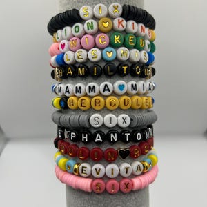 May include: A collection of colourful beaded bracelets. Each bracelet has letter beads spelling out musical titles including "SIX", "LION KING", "WICKED", "LES MIS", "HAMILTON", "MAMMA MIA", "HERCULES", "PHANTOM", and "EVITA". The bracelets are in various colours.