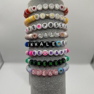 May include: A selection of beaded bracelets with motivational words. The bracelets display words such as "HOPE", "BE KIND", "IS POSSIBLE", "CALM", "INHALE", "BE HAPPY", "STRENGTH", and "DREAM" using white, colourful, and black beads.