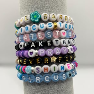 May include: A collection of colourful beaded bracelets. The bracelets are made of white, blue, pink, black, and purple beads. They spell out phrases such as "CIRCUS TOUR" and "NEVER FORGET". Some have star and flower charms.
