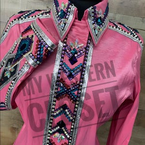 Może przedstawiać: Żyworóżowa koszula westernowa z misternymi zdobieniami. Koszula posiada spiczasty kołnierz i mankiety, ozdobione kolorowymi dżetami, cekinami i srebrnymi wykończeniami. Przód posiada dekoracyjny panel o podobnym wzorze. Koszula z długim rękawem jest prezentowana na manekinie.