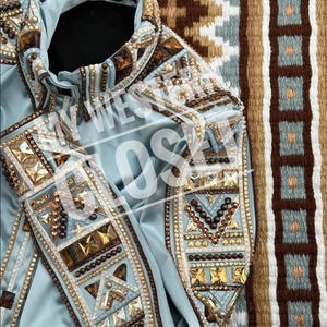 Könnte beinhalten: Hellblaue Showjacke mit aufwendigen Verzierungen. Die Jacke hat einen Kragen und ist mit goldenen, braunen und klaren Strasssteinen und quadratischen Edelsteinen verziert. Die Ärmel und die Vorderteile sind stark mit geometrischen Mustern verziert.