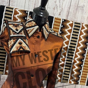 Op de afbeelding: Een bruin paardenshowshirt met decoratieve versieringen op de kraag en schouders. Het shirt wordt getoond op een mannequin met een bijpassend zadeldek op de achtergrond. De tekst "MY WEST CLOS" is zichtbaar op het shirt.