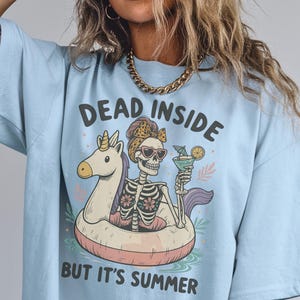 May include: Light blue t-shirt with the text "DEAD INSIDE BUT IT'S SUMMER". The graphic features a skeleton wearing sunglasses and a headscarf, floating on a unicorn-shaped pool float and holding a cocktail.