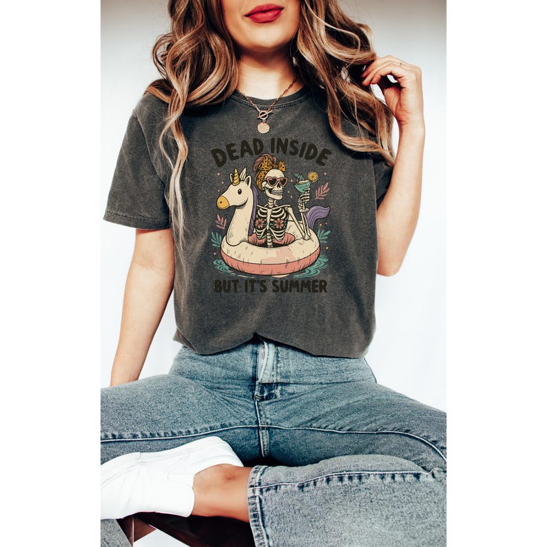 May include: Dark gray t-shirt with the text "DEAD INSIDE BUT IT'S SUMMER". The graphic features a skeleton in a pool floatie with a unicorn head, holding a cocktail. The shirt is worn by a person.