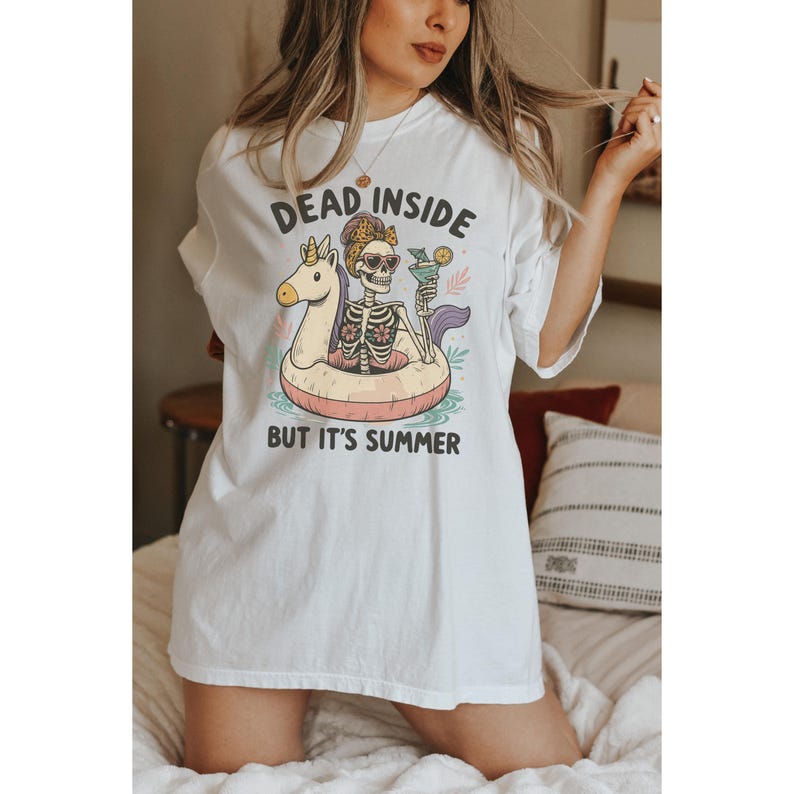 May include: White t-shirt with the text "DEAD INSIDE BUT IT'S SUMMER". The graphic shows a skeleton in sunglasses and a floral headband, floating on a unicorn-shaped pool float with a cocktail. The design includes pastel colors.