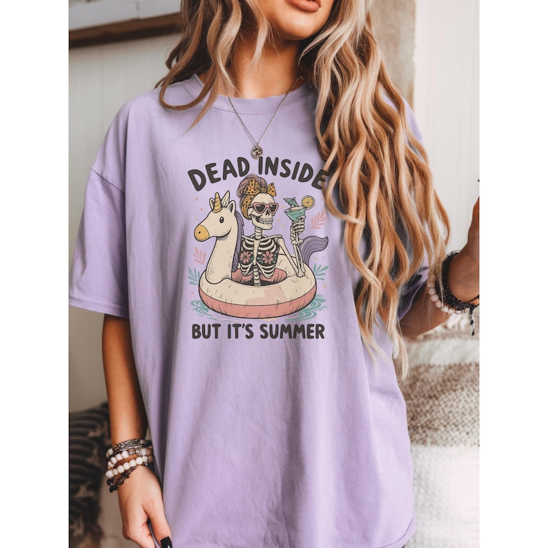 May include: A lilac-colored t-shirt with the text "DEAD INSIDE BUT IT'S SUMMER" above a skeleton graphic. The skeleton is in a pool floatie with a cocktail. The graphic is in black, white, and tan.