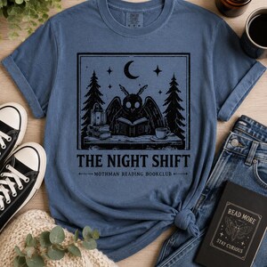 Può includere: Una t-shirt blu a maniche corte con una grafica nera che raffigura una creatura alata intenta a leggere un libro a una scrivania, circondata da alberi, stelle e una luna crescente. Il testo recita THE NIGHT SHIFT MOTHMAN READING BOOKCLUB.