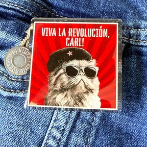 May include: A square, clear acrylic pin featuring a cat wearing sunglasses and a beret, set against a red background with radial lines. The text "VIVA LA REVOLUCIÓN, CARL!" is printed above the cat.