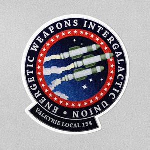 May include: A circular patch with the text "Energetic Weapons Intergalactic Union" and "Valkyrie Local 154". The patch features a blue background with white stars and three rockets with white smoke trails.