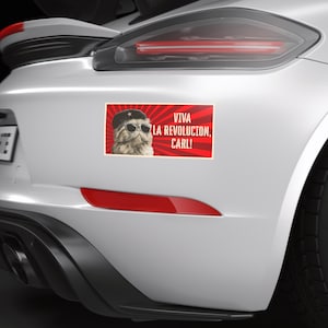 Könnte beinhalten: Rechteckiger Aufkleber auf einem weißen Auto mit einer Katze, die eine Sonnenbrille und eine Baskenmütze trägt. Der Aufkleber hat den Text "VIVA LA REVOLUCION, CARL!" auf rotem Hintergrund mit radialen Linien.