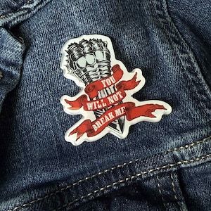 Puede incluir: Un pin transparente troquelado con una ilustración estilizada de puños apretados con una pancarta que dice "YOU WILL NOT BREAK ME" en rojo. El pin está colocado sobre una chaqueta vaquera azul.