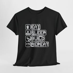 May include: Black t-shirt with the words "EAT SLEEP BUILD REPEAT" in white block letters. The design includes icons of a coffee cup, a bed, a wrench, and a circuit board. The shirt is made of a soft material.