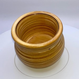 Puede incluir: Un cuenco de madera pequeño y redondo con un acabado liso y pulido, que presenta patrones de grano horizontales visibles en tonos alternos de marrón claro y medio. El cuenco tiene una abertura ancha y un cuerpo abombado con un borde grueso y redondeado.