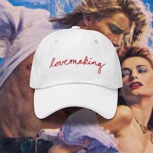 Puede incluir: Gorra de béisbol blanca con la palabra "lovemaking" bordada en escritura roja. La gorra se coloca sobre un fondo de estilo vintage que presenta una escena romántica con dos personas.