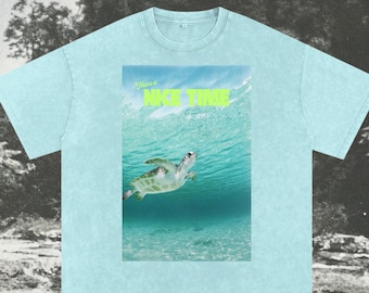 May include: Light blue t-shirt with a graphic of a sea turtle swimming underwater. The image includes the text "Have a NICE TIME" in neon green. The shirt is made of a soft material.