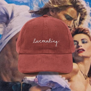 Puede incluir: Gorra de béisbol de pana rojo ladrillo con la palabra "lovemaking" bordada en escritura blanca. La gorra tiene una visera curva y un diseño de seis paneles. El fondo presenta una ilustración vintage.