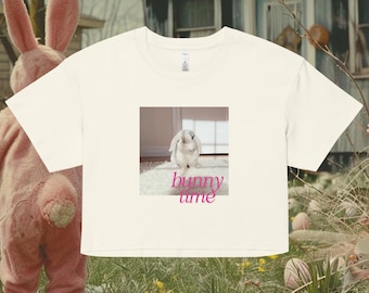 May include: Cream-colored t-shirt featuring a square graphic of a white bunny on a fluffy rug, with the words "bunny time" in pink. A person in a pink bunny costume is in the background.