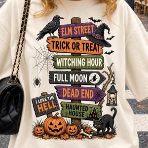 May include: A cream-colored t-shirt featuring a graphic of a wooden signpost with multiple directional signs reading Elm Street, Trick or Treat, Witching Hour, Full Moon, Dead End, Haunted House, and I Love the Hell, with pumpkins, a cat, and a crow.