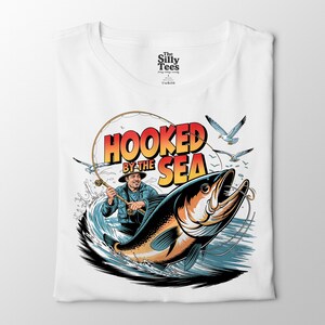以下が含まれることがあります： 白いTシャツ。大きな魚を釣る漁師のグラフィックで、「Hooked by the Sea」の文字がオレンジと黄色で描かれています。デザインにはカモメと水しぶきが含まれています。