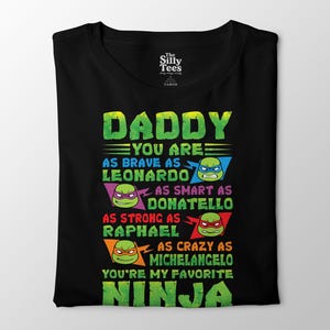 Op de afbeelding: Zwart T-shirt met een groen en gele opdruk met de tekst "DADDY YOU ARE AS BRAVE AS LEONARDO AS SMART AS DONATELLO AS STRONG AS RAPHAEL AS CRAZY AS MICHELANGELO YOU'RE MY FAVORITE NINJA". Het shirt heeft een ronde hals.