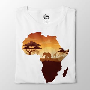 Puede incluir: Camiseta blanca con un diseño del continente africano al atardecer. El diseño incluye siluetas de un elefante, una jirafa y árboles en tonos marrones y naranjas. El texto "The Silly Tees" está impreso en la parte superior.