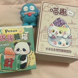 May include: An assortment of colorful collectible boxes and figurines. One box features a panda bear illustration with Chinese characters. Another box displays a cat design with a crystal. A blue figurine with a tongue sticking out is also present.
