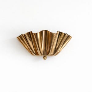 Op de afbeelding: Een goudkleurige wandlamp met een geschulpte, waaiervormige vormgeving. De lamp heeft een klein, rond detail aan de onderkant. De armatuur is gemaakt van metaal en staat tegen een witte achtergrond. Dit decoratieve stuk is ideaal voor de interieurdecoratie.