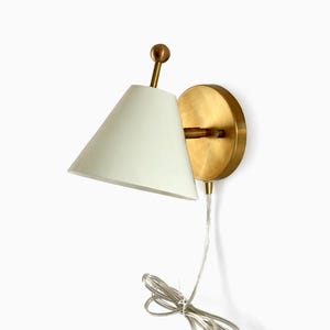 Pode incluir: Luminária de parede com acabamento em latão e abajur cônico creme. A luminária tem uma base redonda e um cabo transparente. O design é minimalista e moderno, adequado para iluminação interior.