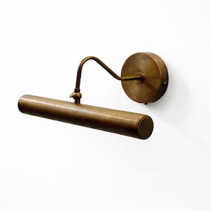 Op de afbeelding: Een messing schilderijlamp in vintage stijl. De lamp heeft een cilindrische behuizing, een gebogen arm en een ronde wandbevestiging. De lamp is bevestigd aan een witte muur.