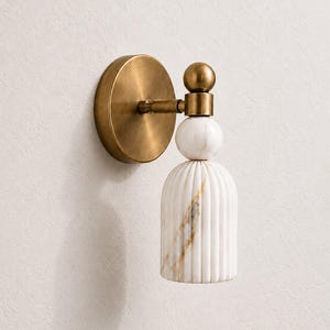 May include: A wall sconce featuring a circular brushed brass backplate and a horizontal arm connected to a brass sphere. Below the sphere is a smooth white marble ball and a fluted white marble cylindrical shade with gold veining throughout.