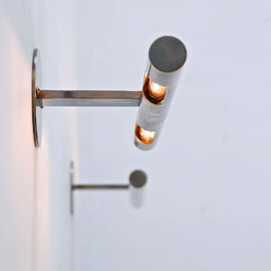 Könnte beinhalten: Zwei moderne, zylindrische Wandleuchten mit warmem Licht, das von innen kommt. Die Leuchten sind aus gebürstetem Metall gefertigt und haben ein minimalistisches Design, das an einer weißen Wand befestigt ist. Ideal für die Raumbeleuchtung.