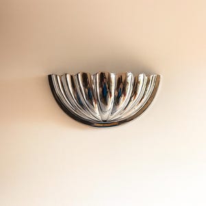 May include: A polished chrome wall sconce with a half-circle shape and a fluted design. The sconce is mounted on a light-coloured wall, reflecting the surrounding environment.