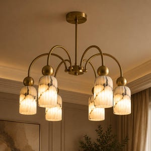 May include: A brass chandelier featuring six curved arms extending from a central rod. Each arm holds a cylindrical, white, and grey marbled glass shade with a smooth, rounded brass connector piece positioned above each light fixture.