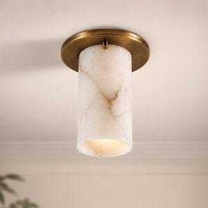 Op de afbeelding: Een plafondlamp met een cilindervormige kap van wit albast met natuurlijke grijze aders. De kap is bevestigd aan een ronde plafondplaat van geborsteld messing die direct op het plafondoppervlak wordt gemonteerd.