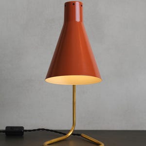 Może przedstawiać: Lampa stołowa w stylu retro z stożkowym, rdzawym kloszem i podstawą z metalu w kolorze mosiądzu. Lampa ma smukłą, zakrzywioną nóżkę i trójkątną podstawę. Przewód i wtyczka są czarne. Lampa stoi na ciemnej powierzchni.