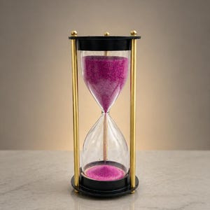 May include: An hourglass with a black top and base, gold-colored metal supports, and clear glass chambers. The sand inside is a vibrant magenta color. The hourglass is a decorative item, perfect for a desk or shelf.