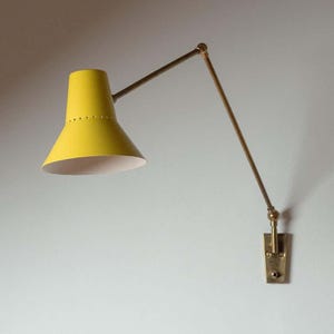 May include: A yellow wall lamp with a conical shade and a brass-coloured adjustable arm. The shade has a white interior and small perforations near the top. The lamp is attached to a brass-coloured rectangular base.