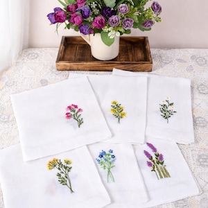 Könnte beinhalten: Weiße Leinentaschentücher mit bunten Blumenmotiven bestickt, darunter rosa, gelbe, blaue und lila Blumen. Ein Strauß lila Blumen steht in einem weißen Krug auf einem Holztablett.