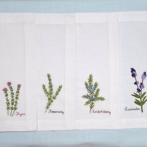 May include: Four white linen napkins with embroidered botanical designs. Each napkin features a different herb: thyme, rosemary, juniper berry, and lavender. The names of the herbs are embroidered below the designs in a cursive font.