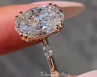 May include: A gold ring featuring a large, oval-cut diamond as the centerpiece. The ring has a delicate band with smaller accent diamonds. The ring is held up, showcasing its sparkle and design. The ring is a piece of jewelry.