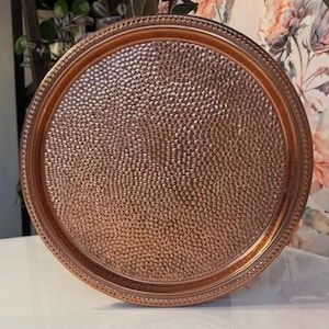 May include: A round copper-coloured tray with a textured surface and a decorative beaded edge. The tray is displayed on a white surface, showcasing its metallic sheen. The tray is approximately 30 cm in diameter.